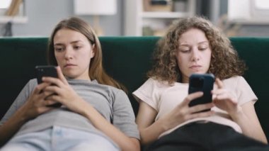 Gündüz akıllı telefon ekranına bakan genç kadınların telefon bağımlılığı. Kanepede otururken, yorgun görünüyorlar, birbirlerini görmezden geliyorlar.