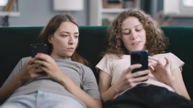 İki neşeli kadın, sosyal medyada yayınlamak için akıllı telefona sarılıyor ya da birbirlerini gösteriyorlar, mutlu arkadaşlar rahatça kanepede dinleniyorlar.