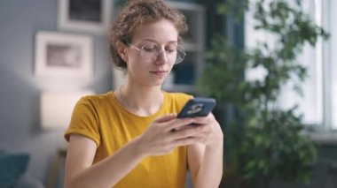 Kıvırcık genç kadın akıllı telefondan hoşlanıyor, gülümseyerek internet mesajlarına bakıyor, mobil iletişim teknolojisinin rahatlığının tadını çıkarıyor.