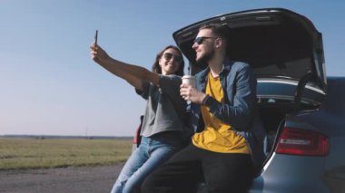 Neşeli çift arabanın bagajında oturur, termostan kahve ya da çayın tadını çıkarırlar, telefonda gülümseyerek selfie çekerler, hepsi güneş ışığında güneşlenirler.
