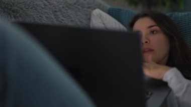 Güzel genç bir kadın yastıkla rahat bir kanepeye uzanıyor, ev kıyafetleri giyiyor, bilgisayardan ilginç bilgiler izliyor ve gülümsüyor.