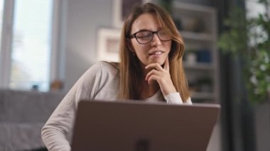 Gözlüklü genç, neşeli bir kadın arkadaşlarıyla görüntülü konuşma yapmak için dizüstü bilgisayar kullanıyor. Sıcak güneşli oturma odasındaki kanepenin yanında oturuyor.