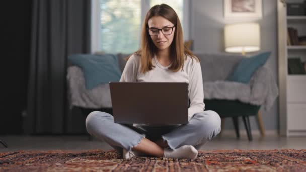 迷人的年轻女子坐在地板上 被笔记本电脑屏幕 浏览和取悦所吸引 穿着舒适的家居服 — 图库视频影像
