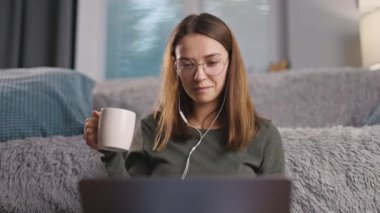 Kulaklıklı genç profesyonel bir kadın evde çalışıyor, dizüstü bilgisayar kullanıyor, kahve içiyor ve keyif alıyor, serbest çalışan birinin yaşam tarzı.