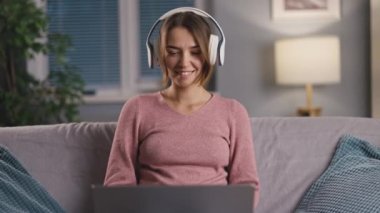 Pembe kazak giyen, kulaklık takan ve dizüstü bilgisayarda müzik dinleyen neşeli gülümseyen kadın, en sevdiği sonbahar çalma listesini yapıyor.