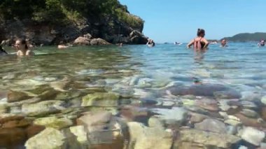 Bir grup insan plajın berrak sularında ferahlatıcı bir yüzüşün tadını çıkarıyor. Güneş, onlar sığ sularda sıçrayıp oynarken parlak bir şekilde tepemizde parlıyor ve neşeli bir yaz sahnesi yaratıyor..