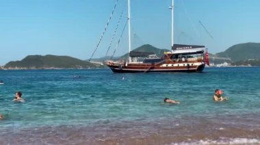 Adriyatik Denizi 'nde güzel bir gün. İnsanlar kristal berrak suda yüzerken büyük bir ahşap tekne kıyıdan geçiyor. Resimli manzara, dağlık bir zemin ve içinde küçük bir ada içerir.