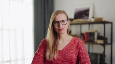 Sarışın, sevimli ve olgun bir bayan. Evde gözlüğünü çıkartıyor. Modern iç dekorasyonlu oturma odasının bulanık arka planı. Mutlu kadın portresi.