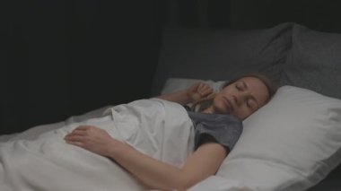 Sarı saçlı, gri pijamalı yumuşak yastıklar ve battaniyeli rahat bir yatakta dinlenen bir kadın. Kadın geceleri huzurlu bir uyku çekiyor..