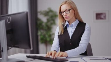 Orta yaşlı, gözlüklü, resmi giyinen beyaz bir kadın evdeki iş yerinde otururken bilgisayar başında daktilo kullanıyor. Kadın serbest çalışan, uzaktan kumandayla çalışıyor..