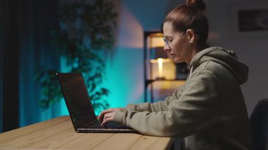 Masada oturan ve modern dizüstü bilgisayarda yazan gözlüklü ciddi bir kadının yan görüntüsü. Akşam saatlerinde evde kadın serbest kodlama verisi.