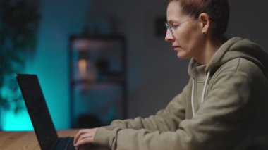 Kadın ağ geliştirici karanlık atmosferli oturma odasında oturuyor ve modern kablosuz dizüstü bilgisayarda yazıyor. Gözlüklü kadının evden geç saatlere kadar çalışmasının yan görüntüsü.