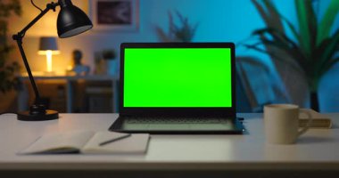 Krom ekranlı modern dizüstü bilgisayar. Masanın üzerinde bir fincan sıcak kahve, notlar ve lamba duruyor. Koyu oturma odasında. Yaklaş, yakınlaş..