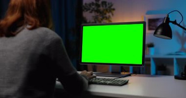 Akşam saatlerinde oturma odasında oturan ve krom ekranlı modern bilgisayarı uzaktan çalışma için kullanan bir kadının arka görüntüsü. İnsan kavramı, serbest meslek ve teknoloji.