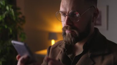 Karanlık odada otururken akıllı telefon kullanan sakallı, yetişkin bir adam. Modern aletlerle evde vakit geçiren beyaz bir erkek..