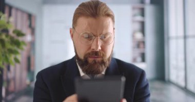 Ofis masasında otururken dijital tablet kullanan kendinden emin bir iş adamı. Beyaz sakallı adam şık bir takım elbise ve kravat giyiyor..