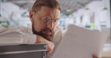 Kafkasyalı işadamı ofiste sıkı çalışırken bilgisayar monitörüne yaslanıyor ve belgeleri okuyor. Resmi giyinmiş olgun bir adam aşırı çalışmaktan yorgun ve uykulu hissediyor..