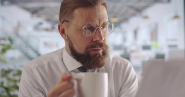 Resmi giyinmiş, gözlüklü kahve içen ve ofiste evrakları inceleyen odaklanmış sakallı bir adam. İnsan, iş ve iş kavramı.