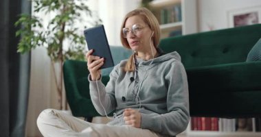 Kulaklıklı orta yaşlı bir kadın kanepenin yanında oturuyor ve dijital tablette görüntülü konuşma yapıyor. Uzak iletişim ve modern yaşam tarzları kavramı.