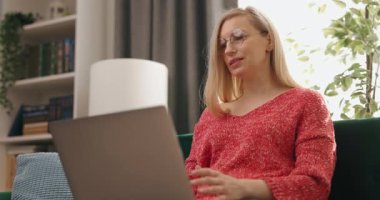 Kanepede oturan ve taşınabilir laptopta görüntülü konuşma yapan gündelik kıyafetli olgun bir kadın. Oturma odasında online sohbet için internete bağlanan serbest kadın..