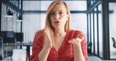 Gözlüklü ve günlük kıyafetleriyle modern ofiste oturan, konuşan ve el kol hareketi yapan odaklanmış beyaz bir kadın. Birinci şahıs görüşü. Video çağrısı kavramı.