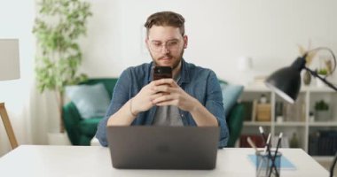 Genç sakallı adam kablosuz bilgisayarla masada otururken akıllı telefondan mesaj atıyor. Evde modern aletler kullanan günlük giysili beyaz bir adam..