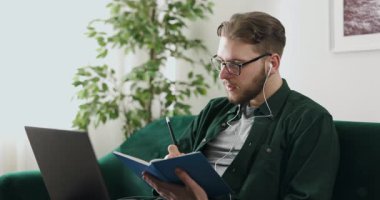 Kablosuz kulaklıklı yakışıklı genç adam evde online çalışmak için modern dizüstü bilgisayar kullanıyor. Odaklanmış erkek öğrenci rahat koltukta oturuyor ve notlar alıyor..