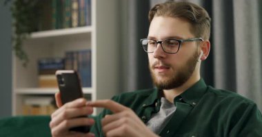 Dijital kulaklıklı genç beyaz bir adam parlak bir dairede koltukta otururken akıllı telefondan müzik listesi çeviriyor. Boş zaman kavramı..