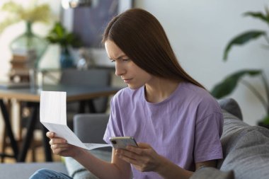 Elinde fatura tutan genç beyaz kadın ve evinde kanepede oturan ödemeyi hesaplıyor. Aile bütçesi, vergi ve faturalar, e-bankacılık konsepti
