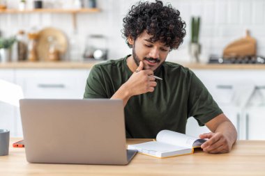 Uzaktan öğrenme, online eğitim. Gülümseyen genç erkek serbest yazar ya da web sitesi izleyen öğrenci portresi ya da evde oturan bir not defterine notlar yazar. Dizüstü bilgisayar kullanan Asyalı adam