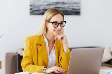 Bilgisayarla çalışan genç ve güzel bir kadın konsantre olmuş ve gülümsüyor modern ya da ev ofisinde oturuyor, uzaktan kumandalı internet işi yapıyor, gülümsüyor.