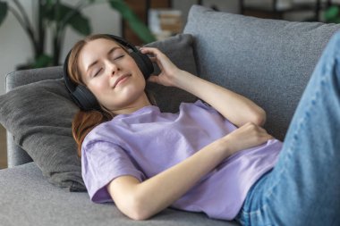 Evde rahatla, olumlu düşün. Mutlu genç bir kadın evinde, kulaklıklarıyla kanepede uzanıyor en sevdiği müziği dinliyor ve güzel bir şey düşünüyor, gülümsüyor.