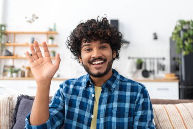 Kıvırcık saçlı Hintli adamın portresi internette sohbet etmek için dizüstü bilgisayar kamerası kullanıyor, video çağrı konsepti, selamlar, kameraya bakıyor ve gülümsüyor.
