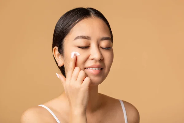 Beautiful korean woman applying a anti age or moisturizer face cream on her face, smiling sweetly standing on a beige background