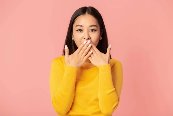 Surprised woman covering her mouth with hands while standing on pink background, looking at the camera