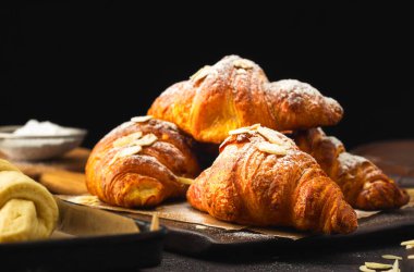 Fotokopi alanı, lezzetli kahvaltı ya da tatlı içeren koyu bir zemin üzerine pudra şekeri ve badem serpiştirilmiş altın çikolatalı kruvasanlar.