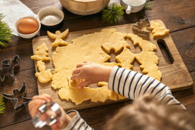 Çocuğun elleri, Noel kurabiyelerini kurabiye keskisi ve hamurla ev manzaralı tahta bir tahtanın üzerinde hazırlıyor. Sıcak tatil pişirme anı