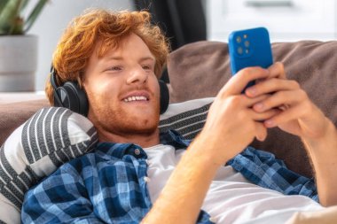 Mutlu, genç, kızıl saçlı bir adam, akıllı telefonuna bakıp, kablosuz kulaklıkla müzik dinlerken kanepede rahatlar. Görüntü, boş zamanları, teknolojiyi ve çevrimiçi eğlenceyi temsil eder.
