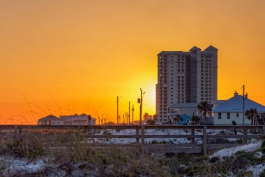 GULF SHORES, AL, USA - APRIL 2, 2021: Brilliant sunset on the beach with sand dunes, buildings and an unidentifiable person crossing a boardwalk clipart