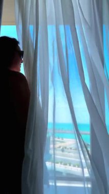 Genç bir kadın açık pencereyle yatakta oturuyor ve deniz manzarasına bakıyor.