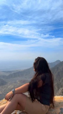 Genç bir kadın dağın tepesinde oturuyor ve arka plandaki dağın tepesinden bir manzarayla ufka bakıyor.