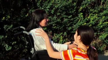 Genç bayan bakıcı, yaşlı bir kadını tekerlekli sandalyeyle şehir caddesinde gezdirirken ve yaz günü yürürken onunla sohbet ederken. Tekerlekli sandalyedeki engelli büyükanneyle ilgilenen genç kız, yaşlı nesle bakıyor.. 