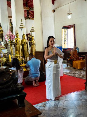 Bangkok, Tayland 'daki Wat Arun Tapınağı' nda ruhani bir bağlılık anı. Geleneksel Tayland kıyafetleri içinde dua eden bir bayan, bu kutsal ve huzurlu yerde dualarını ve meditasyonlarını sunan Budist bir keşiş ve diğer dindarların yanında diz çöker. 