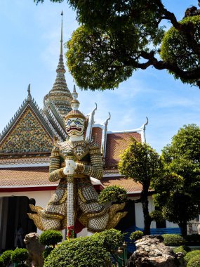 Bangkok, Tayland 'daki Wat Arun Tapınağı' nda yemyeşil ağaçlar ve parlak mavi gökyüzü tarafından çerçevelenmiş renkli dev bir iblis koruyucu heykel. Heykel, heybetli doğal ortamda görkemli bir şekilde dururken bir güç ve koruma havası yayar.