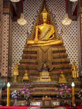Tayland, Bangkok 'taki Wat Arun Tapınağı' nda renkli çiçeklerle bezenmiş bir tahtta oturan büyüleyici bir Altın Buda heykeli. Heykelin ve tapınağın dekoratif mimarisinin karmaşık ayrıntıları büyüleyici bir ortam yaratıyor.
