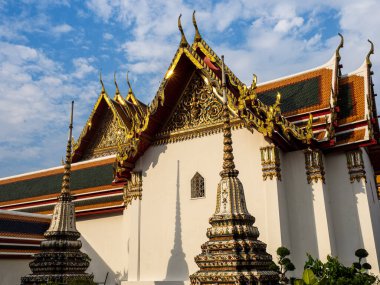 Wat Pho Tapınağı 'nın bir bölümü. Karmaşık altın çatılı. Güneş ışınlarını yansıtıyor. Duvarda gölgeli iki küçük kule görünüyor. Beyaz bulutlu mavi gökyüzü huzurlu bir zemin oluşturarak tapınağın güzelliğini ve ihtişamını arttırır.