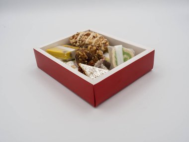 Kırmızı kenarlı bir kutunun açılı görüntüsü çeşitli Hint ghee tatlılarıyla dolu, beyaz arka planda izole edilmiş..