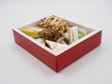 Kırmızı kenarlı bir kutunun açılı görüntüsü çeşitli Hint ghee tatlılarıyla dolu, beyaz arka planda izole edilmiş..