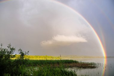 İsveç 'in Hssleholm yakınlarındaki büyüleyici atmosferde, Finjasjn Gölü dramatik bir manzaraya tanıklık ediyor. Huzurlu yüzeyinde dans eden canlı bir gökkuşağı, dramatik bulutların etkileşimi ve ağaçların üzerindeki sıcak güneş ışığının dokunuşuyla çerçevelenmiş..