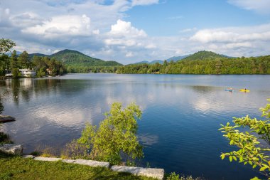 New York 'un kuzeyindeki Sakin Ayna Gölü' nde sakin su üzerinde parlak mavi ve sarı kanolar ve arka planda çarpıcı mavi gökyüzü ve görkemli dağlar bulunur..